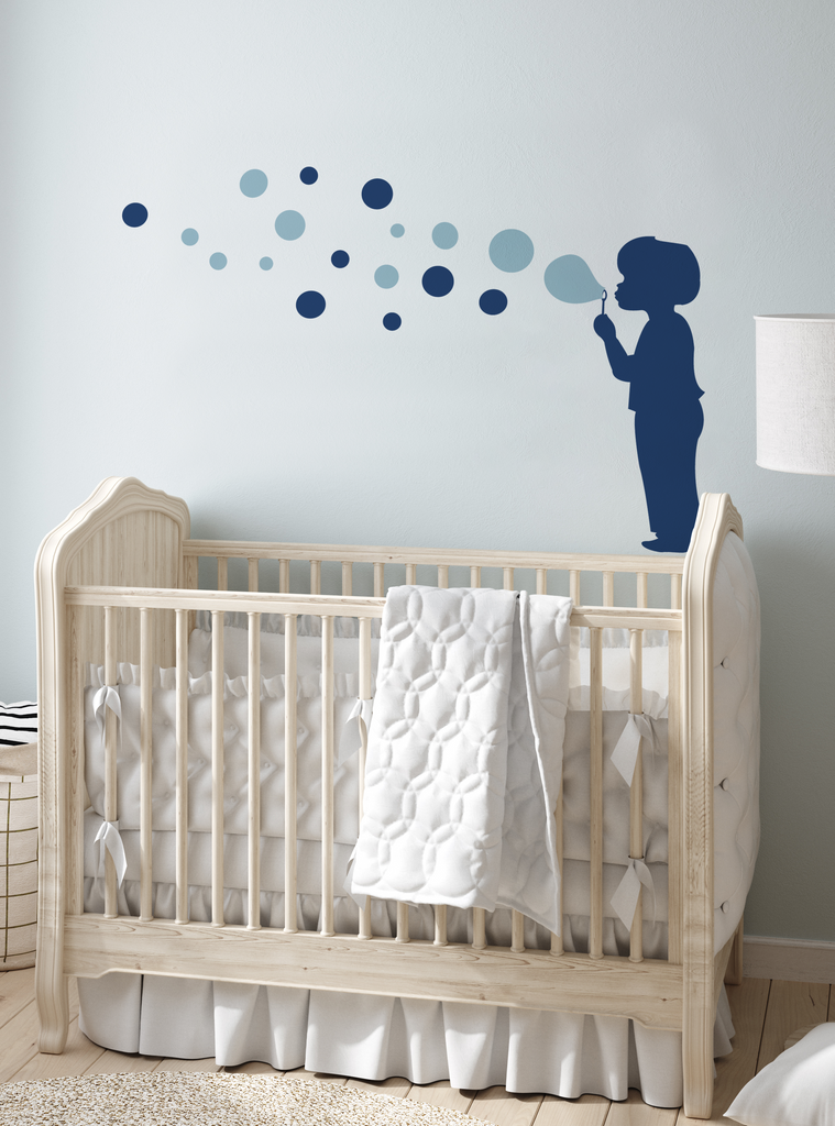 Bubble Boy Decal - Children's Vinyl Wall Decal showcasing a navy blue silhouette of a child blowing bubbles across a light blue wall. The playful design features floating bubbles in varying sizes and shades of blue, displayed above a classic wooden crib with white bedding. Perfect for nursery or children's room decor. Wall Decals, All Products