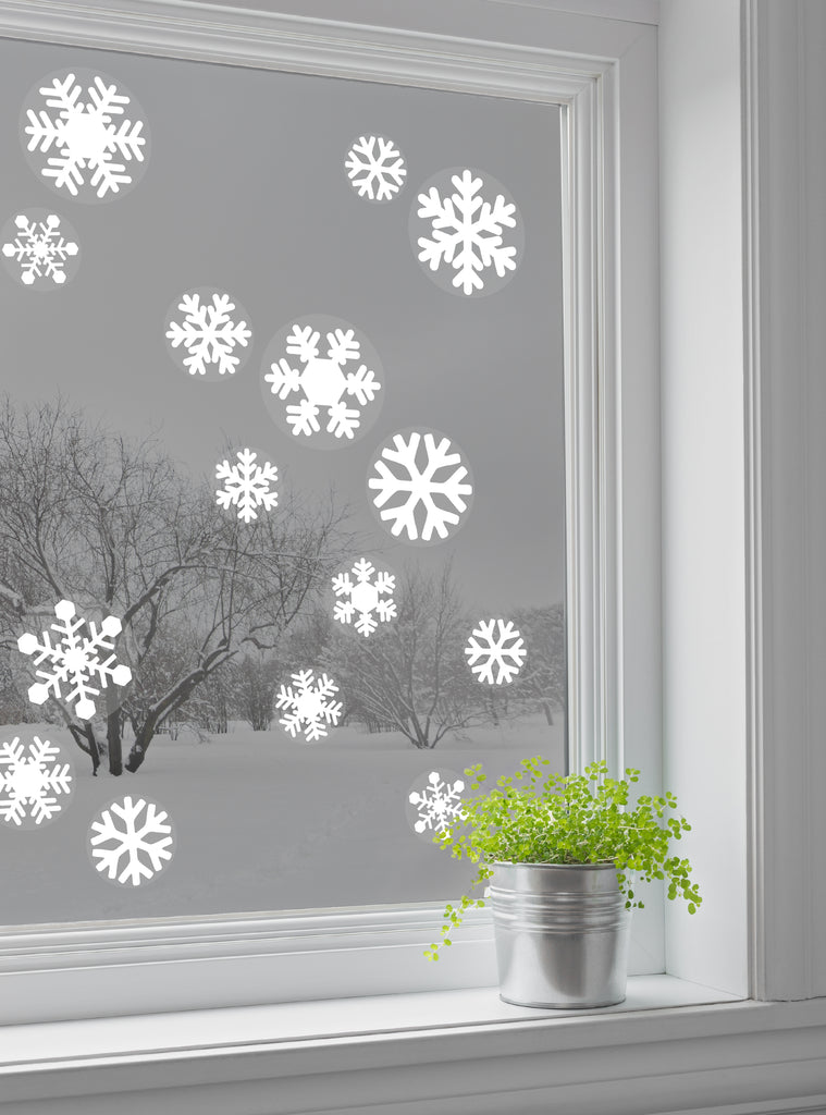 Snowflake Window Stickers displayed on a window, creating a festive winter scene. Various sizes of white snowflake decals contrast beautifully against the gray outdoor view. A small green plant in a silver pot sits on the windowsill, adding a pop of color. Perfect for holiday decorating. Snowflake Window Stickers, Top Sellers, Wall Stickers, Holiday Decals and Stickers, All Products