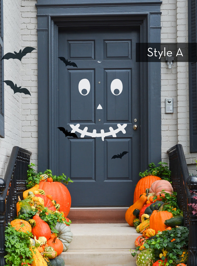 Halloween Pumpkin Face Door Decorations - Vinyl Wall Decal displayed on a charcoal gray door with white facial features creating a playful monster expression. Black bat decals flutter nearby, while vibrant orange pumpkins and greenery adorn the entrance steps, showcasing a festive fall door display. Featured in Wall Decals, Holiday Decals and Stickers, All Products.