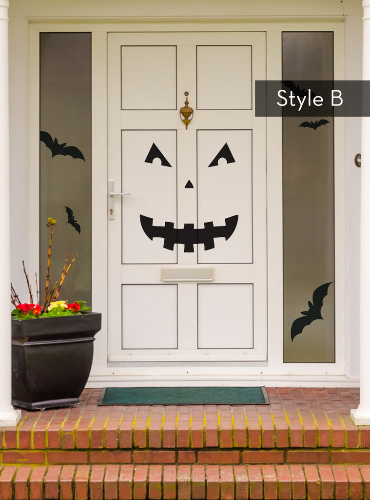 Halloween Pumpkin Face Door Decorations - Vinyl Wall Decal showcased on a white front door displaying Style B face design with triangular eyes, small nose, and jagged smiling mouth. Black bat decorations flutter on the sides, complemented by a decorative planter with autumn foliage on brick steps. Holiday Decals and Stickers, Wall Decals, All Products.