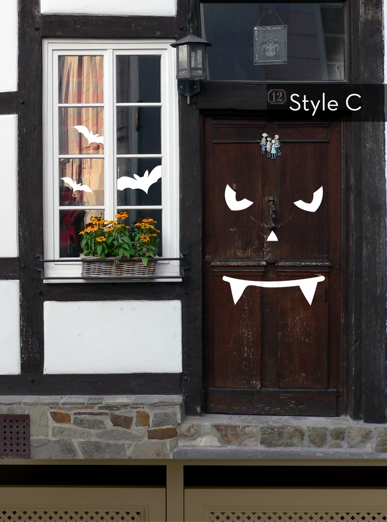 Halloween Pumpkin Face Door Decorations - Vinyl Wall Decal shown on dark wooden door with spooky vampire-style face design featuring angular eyes and fanged mouth. Window beside displays white bat decorations and orange flowers in window box. Traditional timber-framed house exterior setting.