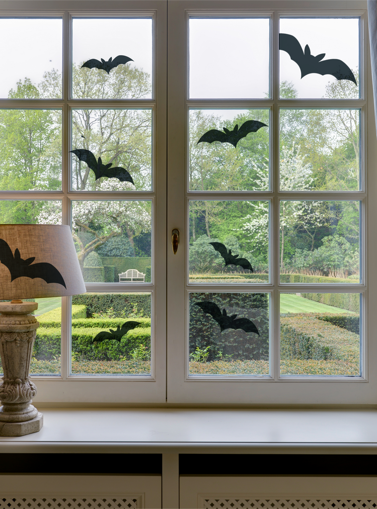 Spooky Bats Wall Decal displayed on a bright window pane overlooking a garden, featuring multiple black bat silhouettes arranged artistically across glass panels. A decorative table lamp sits on the windowsill, creating a festive Halloween atmosphere. Wall Decals, Holiday Decals and Stickers.