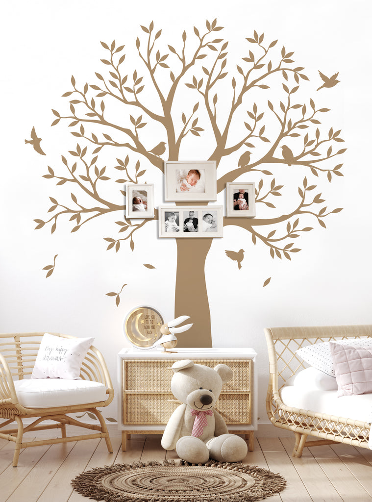 Large brown family tree wall decal with branches and leaves, displaying framed family photos. Decorative birds and falling leaves add charm. Shown in a cozy nursery setting with wicker furniture and a teddy bear. Perfect for showcasing family memories and adding warmth to any room. Top Sellers,Wall Decals,Family Tree Decals,All Products