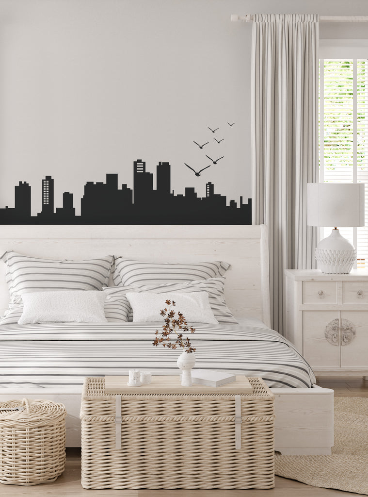 City Skyline Wall Decal shown in a modern bedroom setting featuring a sleek black silhouette of urban buildings and flying birds against a crisp white wall. The large-scale decal creates dramatic contrast above a contemporary bed with striped bedding and wicker storage furniture, Wall Decals, Top Sellers, All Products