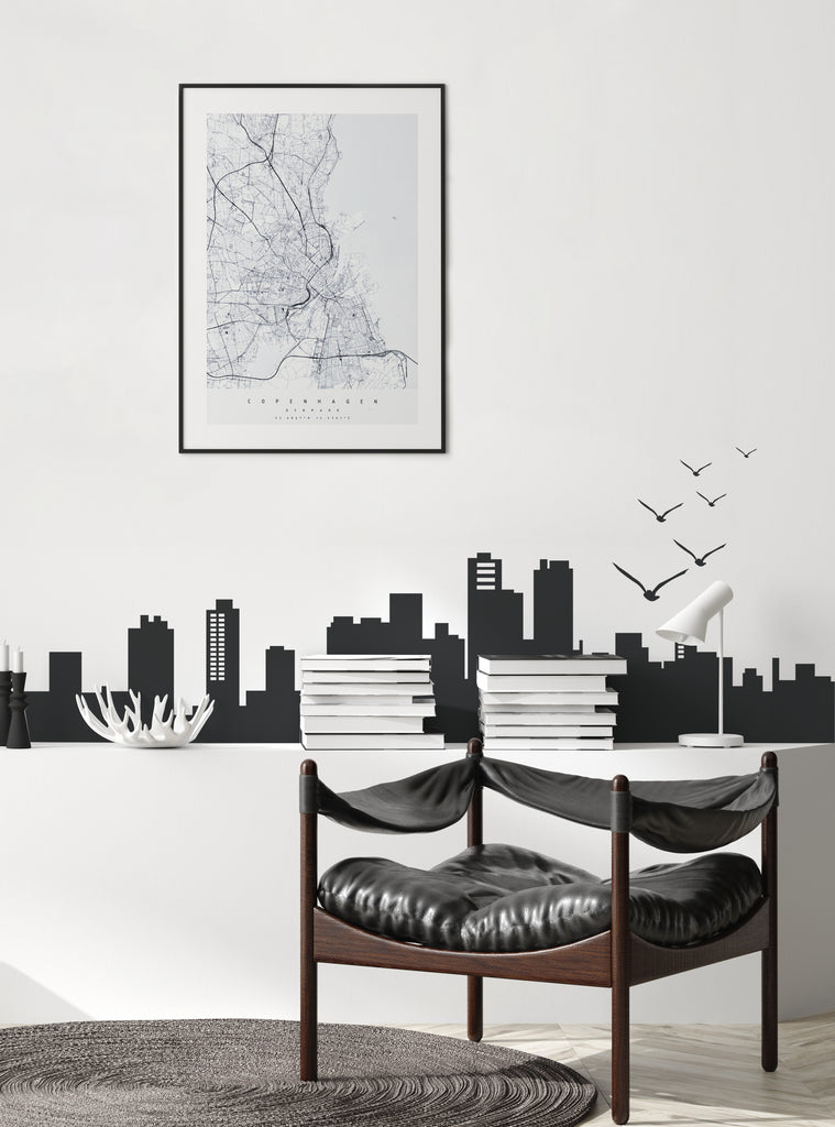 City Skyline Wall Decal displayed in a modern minimalist setting featuring a black silhouette of urban high-rises and buildings spanning across a white wall, with decorative flying birds. Shown above stacked books beside a contemporary leather chair and framed city map artwork.